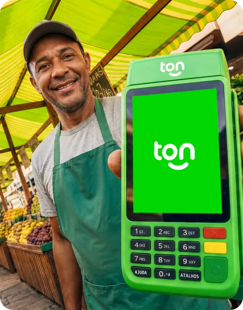 Feirante de avental sorrindo e mostrando a maquininha T3 na banca de frutas